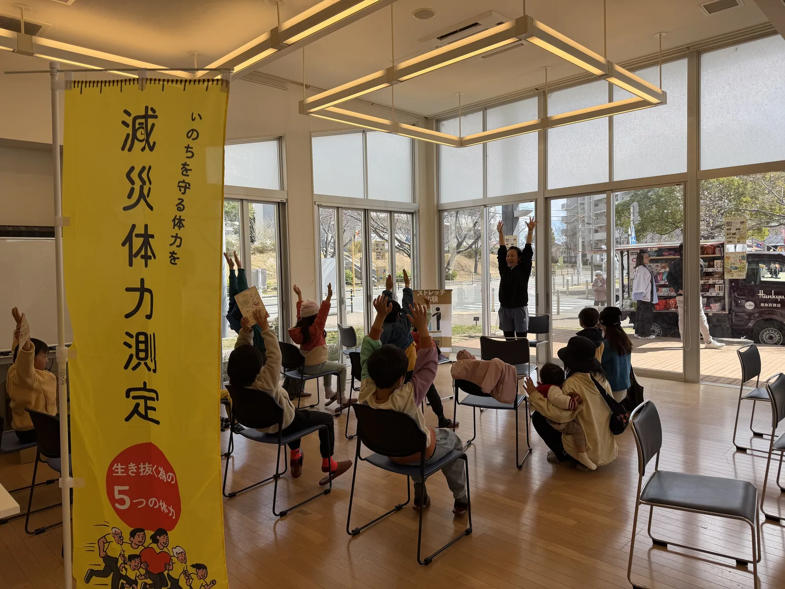 明るい会場で「減災体力測定」ののぼりを背に、参加者が両腕を上げて運動に取り組む様子