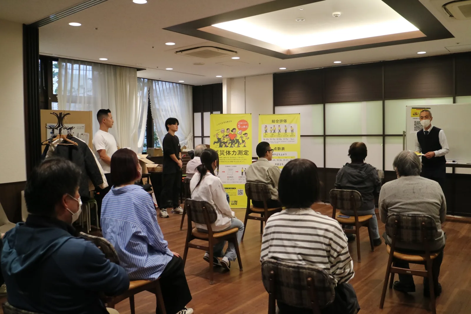 山吹の里での減災体力測定。スタッフが評価ポスターの前で参加者に進め方を説明する様子